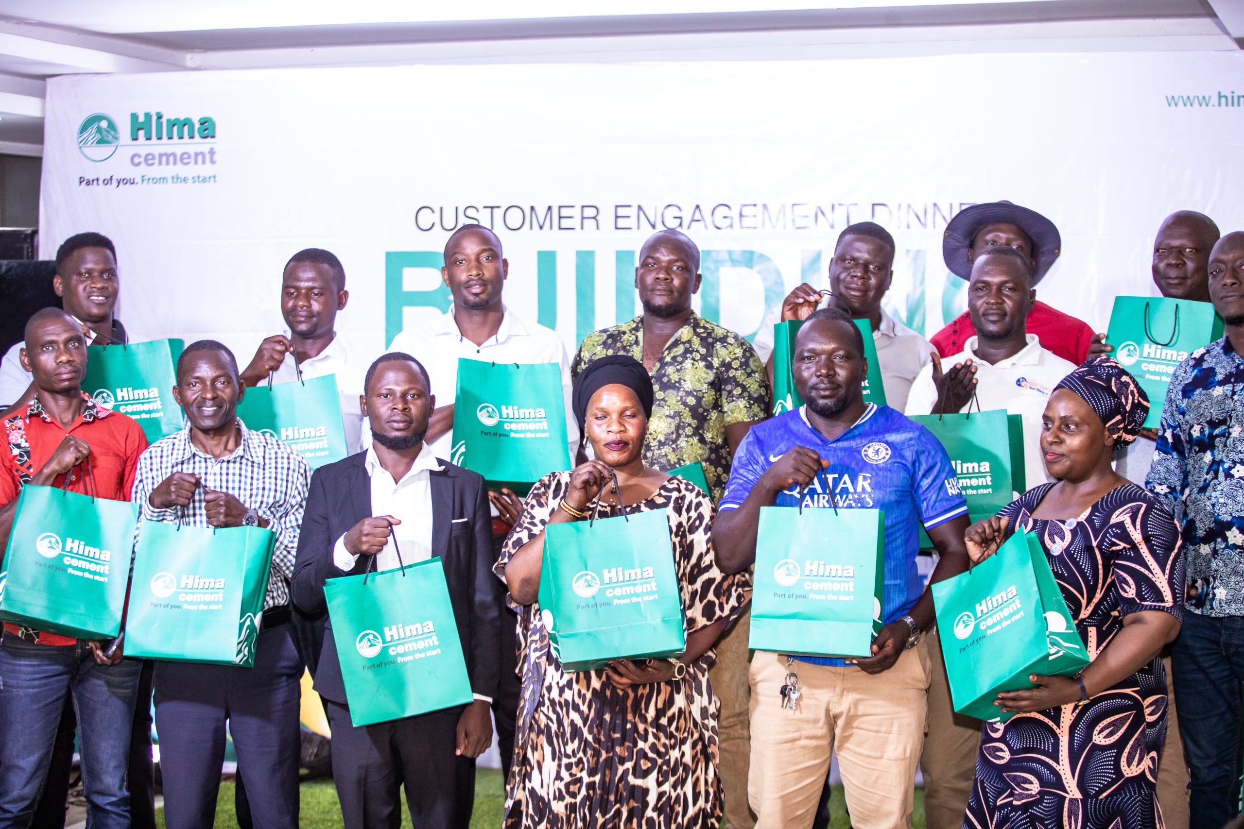 Attendees hold green Hima Cement gift bags while posing in front of a branded customer engagement backdrop in Lira.